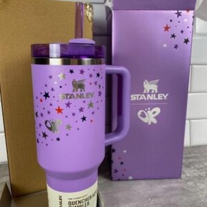 Op de afbeelding: Een lavendelkleurige Stanley Quencher H2.0 FlowState-beker met een handvat, rietje en deksel. De beker is versierd met zilveren vlinders en sterren. De doos is lavendelkleurig met het Stanley-logo en vlindergraphics.