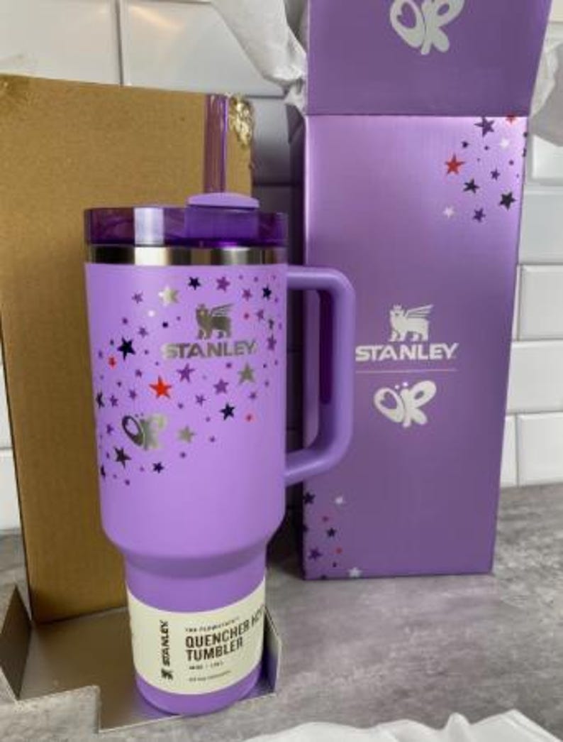 Op de afbeelding: Een lavendelkleurige Stanley Quencher H2.0 FlowState-beker met een handvat, rietje en deksel. De beker is versierd met zilveren vlinders en sterren. De doos is lavendelkleurig met het Stanley-logo en vlindergraphics.