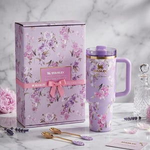 May include: A lavender Stanley Loveshackfancy tumbler with a handle, floral design, and gold accents, next to a matching gift box tied with a pink ribbon. Two gold spoons and a card are also present.