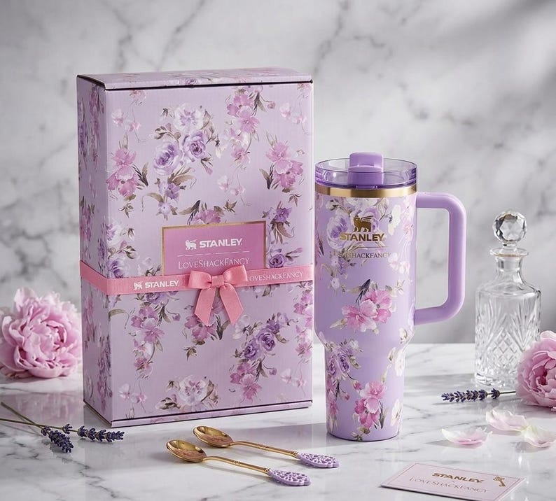 May include: A lavender Stanley Loveshackfancy tumbler with a handle, floral design, and gold accents, next to a matching gift box tied with a pink ribbon. Two gold spoons and a card are also present.