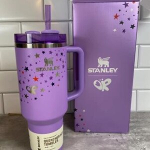 Op de afbeelding: Een lavendelkleurige Stanley Quencher-beker met een handvat, rietje en deksel, naast een bijpassende doos. De beker is versierd met sterren en vlinders. Zowel de doos als de beker zijn voorzien van het Stanley-logo.
