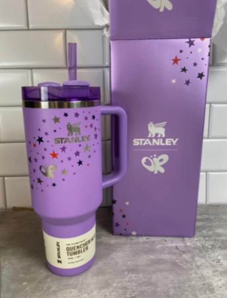 Op de afbeelding: Een lavendelkleurige Stanley Quencher-beker met een handvat, rietje en deksel, naast een bijpassende doos. De beker is versierd met sterren en vlinders. Zowel de doos als de beker zijn voorzien van het Stanley-logo.