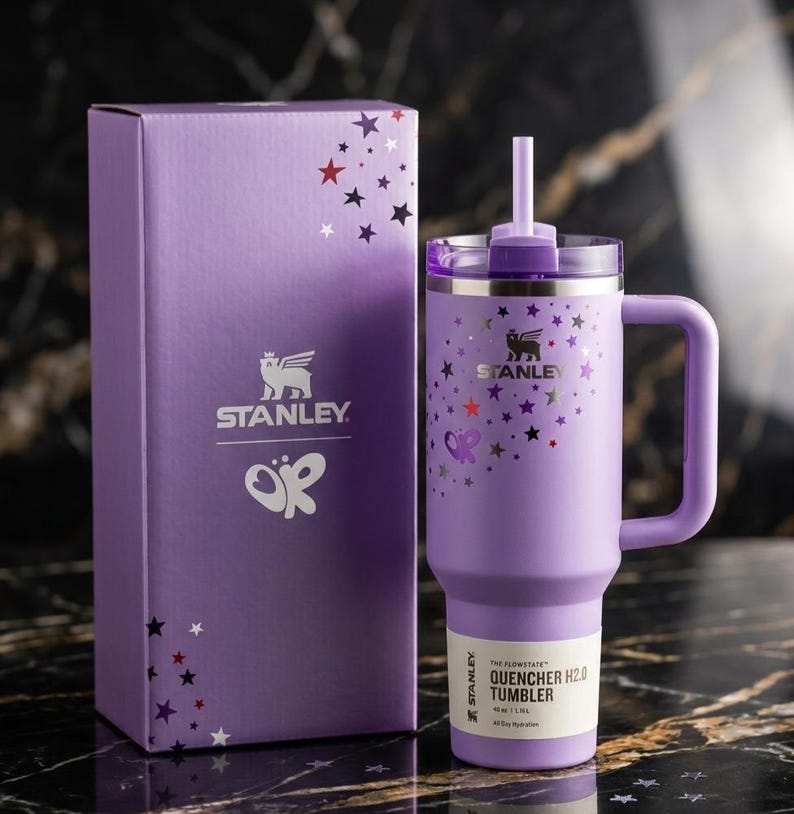 Op de afbeelding: Een paarse Stanley Quencher H2.0 tumbler met een handvat en een rietje, naast een bijpassende doos. De tumbler en de doos zijn versierd met ster- en vlindergraphics. De tumbler heeft het Stanley-logo en de tekst "QUENCHER H2.0 TUMBLER".