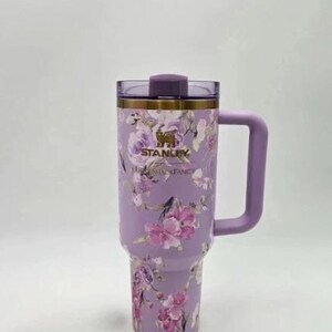Puede incluir: Un vaso Stanley lavanda con un dise&ntilde;o floral con flores rosas y moradas. El vaso tiene un asa, una tapa y el logotipo de Stanley. Las palabras "Stanley" y "Floral Fancy" est&aacute;n impresas en el vaso.