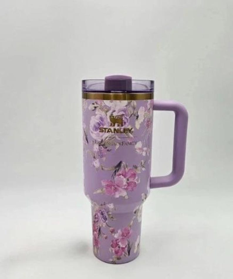 Puede incluir: Un vaso Stanley lavanda con un dise&ntilde;o floral con flores rosas y moradas. El vaso tiene un asa, una tapa y el logotipo de Stanley. Las palabras "Stanley" y "Floral Fancy" est&aacute;n impresas en el vaso.