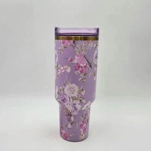 May include: A lavender tumbler with a floral design featuring pink and white flowers. The tumbler has a clear lid and a gold-colored band near the top. The tumbler is designed to hold cold or hot beverages.