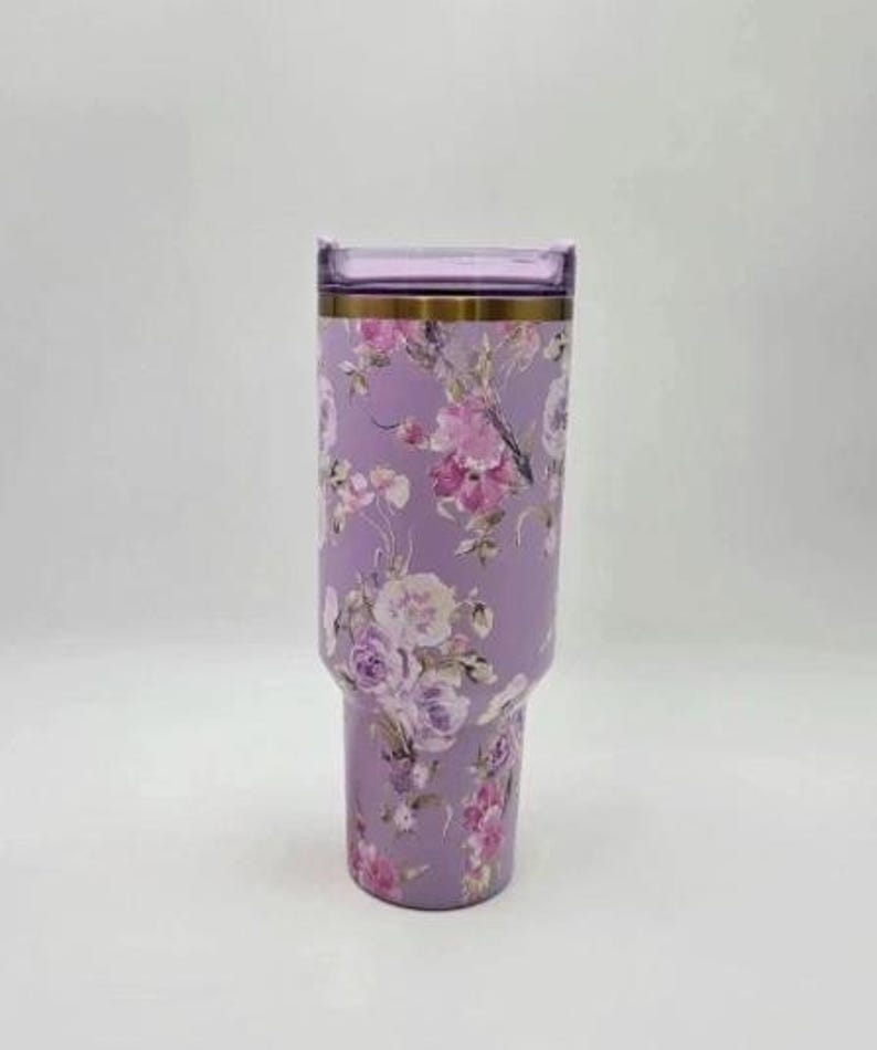 May include: A lavender tumbler with a floral design featuring pink and white flowers. The tumbler has a clear lid and a gold-colored band near the top. The tumbler is designed to hold cold or hot beverages.