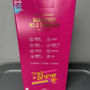 K&ouml;nnte beinhalten: Eine leuchtend pinke Box f&uuml;r einen "QUENCHER H2.0 TUMBLER". Die Box hat goldfarbene Schrift und Symbole, mit dem Text "Time to Shine" und "Barbie 65". Der Becher fasst 1,18 L und h&auml;lt Getr&auml;nke 11 Stunden kalt.