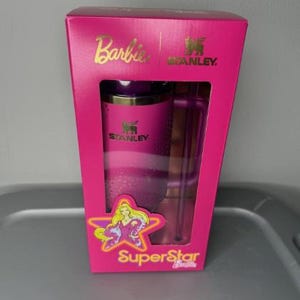 K&ouml;nnte beinhalten: Ein pinkfarbenes Geschenkset von Stanley und Barbie in einer Box. Die Box zeigt das Barbie-Logo, das Stanley-Logo und die Worte "SuperStar". Das Set enth&auml;lt einen pinkfarbenen Becher und einen durchsichtigen Strohhalm.