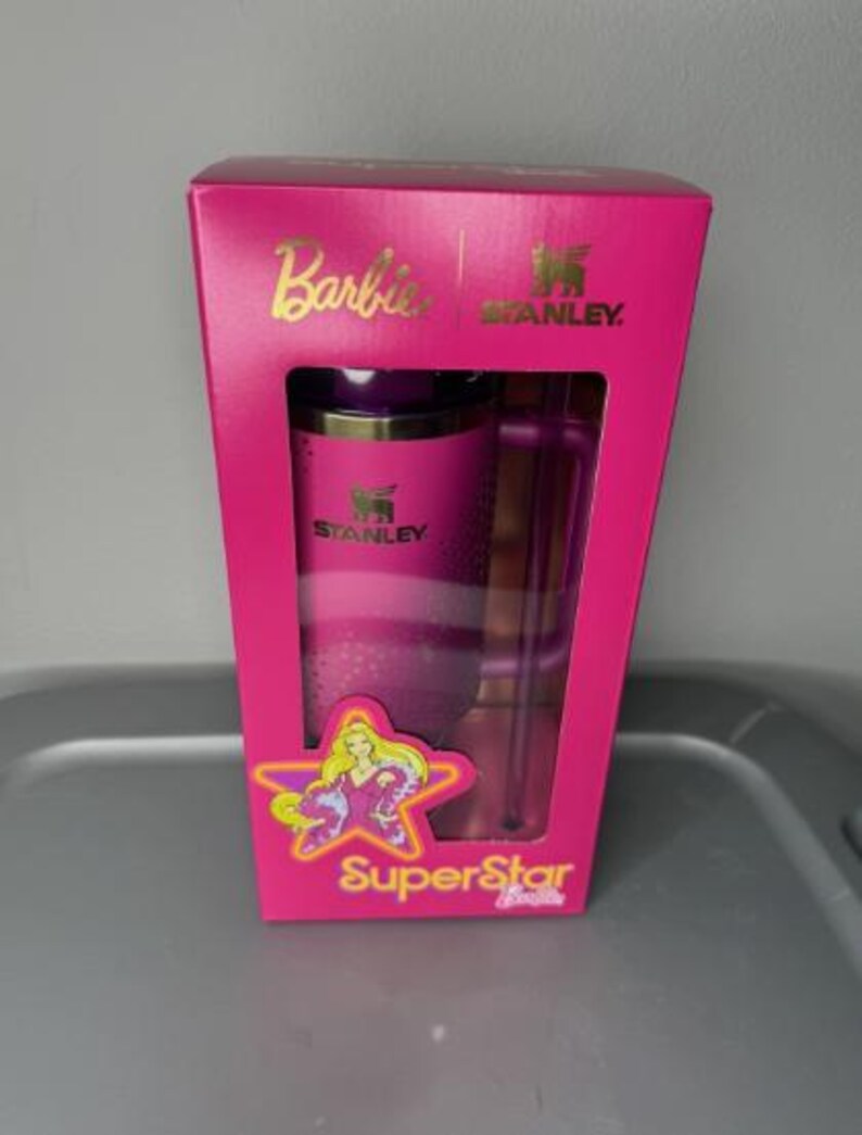 K&ouml;nnte beinhalten: Ein pinkfarbenes Geschenkset von Stanley und Barbie in einer Box. Die Box zeigt das Barbie-Logo, das Stanley-Logo und die Worte "SuperStar". Das Set enth&auml;lt einen pinkfarbenen Becher und einen durchsichtigen Strohhalm.