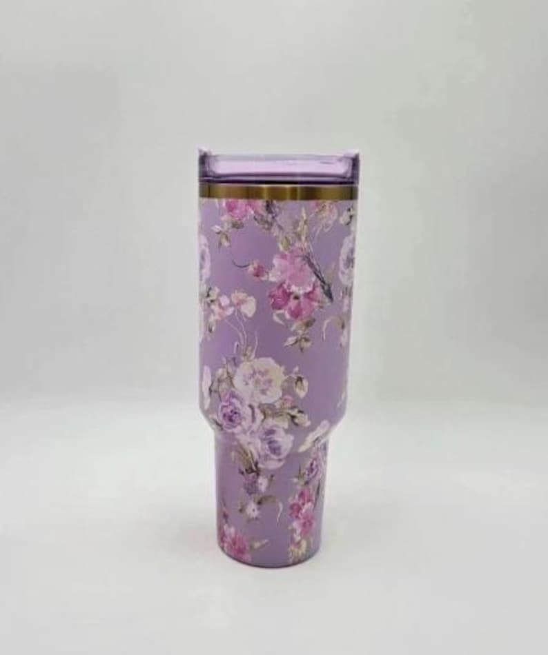 Puede incluir: Un vaso de color lavanda con un dise&ntilde;o floral con flores rosas y blancas. El vaso tiene una tapa transparente y una banda dorada alrededor de la parte superior. El vaso est&aacute; dise&ntilde;ado para contener unos 887 ml.