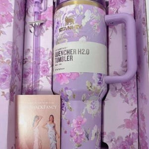 Puede incluir: Un vaso Stanley Quencher H2.0 lavanda con dise&ntilde;o floral, asa y pajita, presentado en una caja a juego. La caja tambi&eacute;n contiene un libro con el texto "SHACKFANCY".