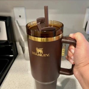 May include: A brown Stanley travel mug with a handle, straw, and a gold band. The mug has a brown lid and is filled with a dark liquid. The Stanley logo is printed in gold on the side of the mug.