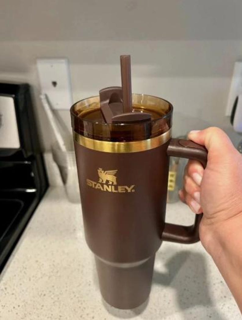 May include: A brown Stanley travel mug with a handle, straw, and a gold band. The mug has a brown lid and is filled with a dark liquid. The Stanley logo is printed in gold on the side of the mug.