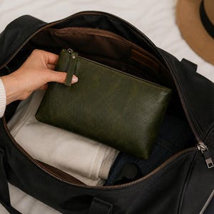 Pu&ograve; includere: Una pochette rettangolare in pelle verde scuro con chiusura a zip, posizionata all'interno di una grande borsa da viaggio nera contenente tessuto bianco piegato e un passaporto blu scuro, con un cappello marrone visibile nell'angolo superiore.