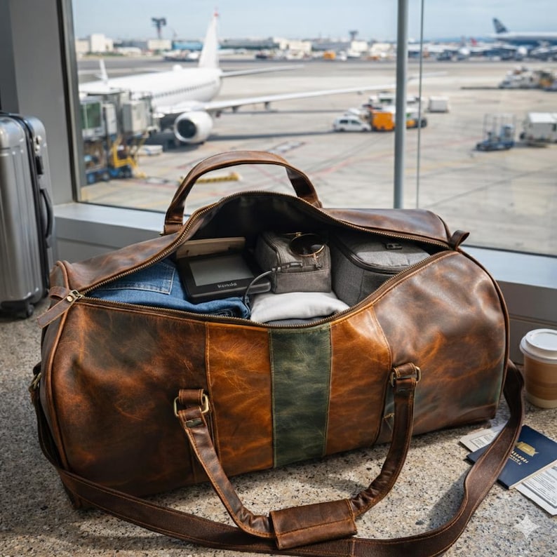 Pu&ograve; includere: Una borsa da viaggio in pelle marrone con una striscia verde al centro, aperta per rivelare oggetti imballati. La borsa ha una tracolla e manici. Lo sfondo mostra un terminal aeroportuale con aerei.