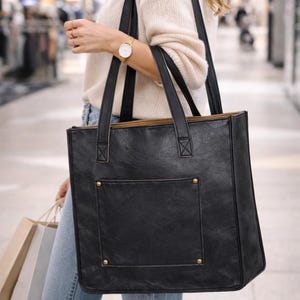 May include: Black tote bag with a rectangular shape, featuring a front pocket and gold-colored rivets. The bag has two black handles and a light brown interior. The bag is carried in a shopping mall.