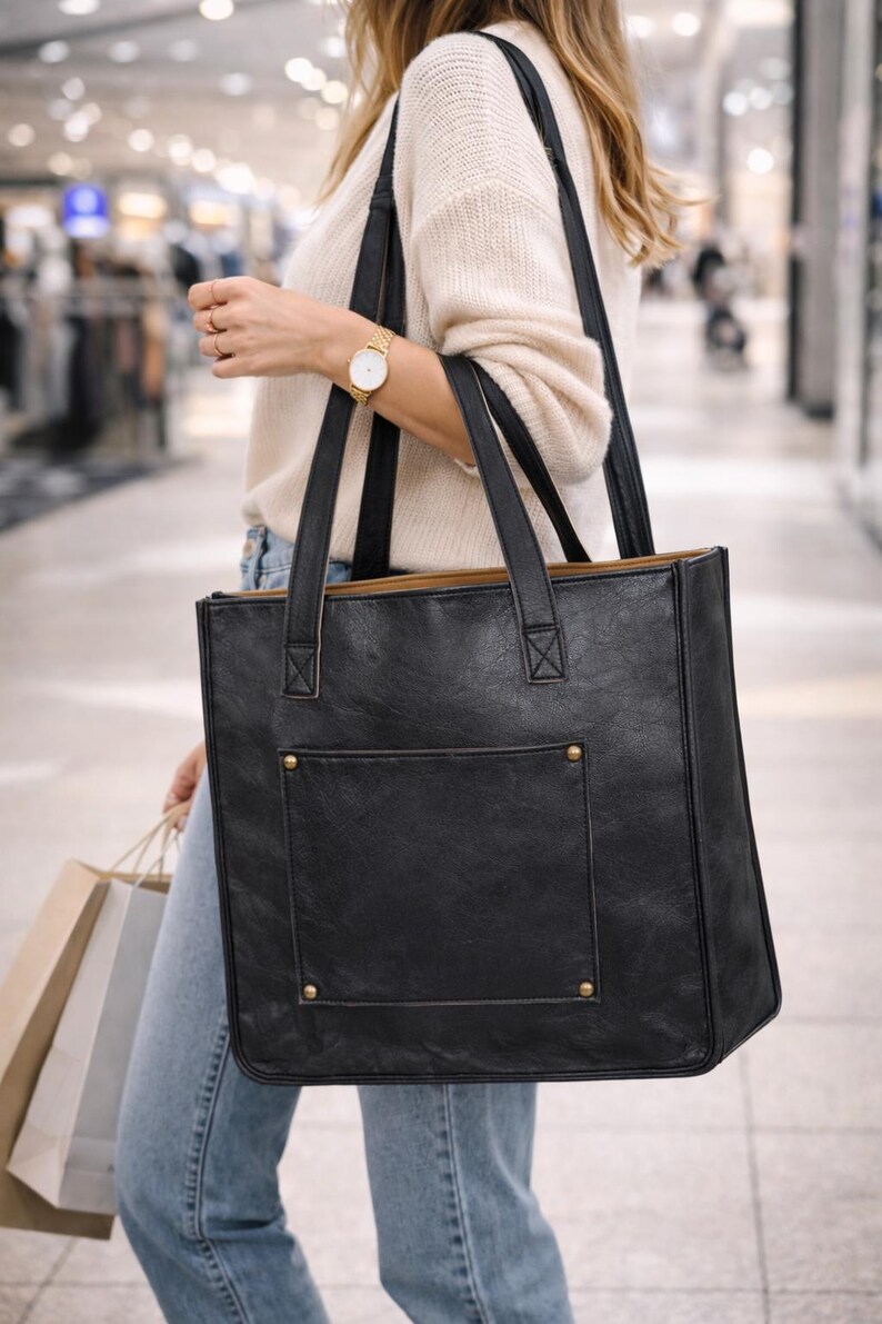 May include: Black tote bag with a rectangular shape, featuring a front pocket and gold-colored rivets. The bag has two black handles and a light brown interior. The bag is carried in a shopping mall.
