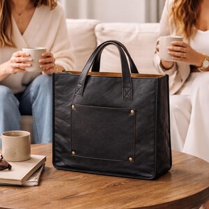 May include: A black leather tote bag with a rectangular shape and a front pocket. The bag has two handles and gold-colored rivets. The interior is a light brown color. The bag is on a wooden table.