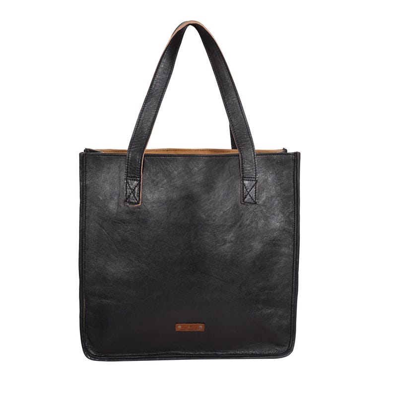 May include: A black leather tote bag with two long handles. The bag has a rectangular shape with a small brown tag on the front. The handles are attached to the bag with reinforced stitching. The interior of the bag is a lighter brown color.