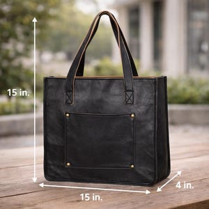 May include: Black leather tote bag with a rectangular shape, measuring 15 inches in height, 15 inches in width, and 4 inches in depth. Features a front pocket, gold-colored rivets, and two top handles. The bag is designed for carrying items.