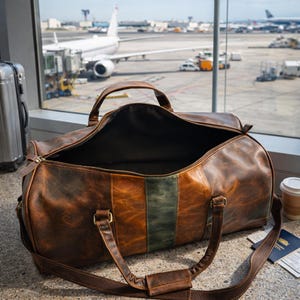 Pu&ograve; includere: Una borsa da viaggio in pelle marrone con una cerniera aperta, che rivela un interno scuro. La borsa ha una striscia verde al centro e si trova vicino a una finestra che si affaccia su un aeroporto con un aereo.