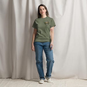 May include: An olive green t-shirt with a circular emblem on the upper left chest. The emblem features a winged skull design with text. The person is wearing blue jeans and white sneakers. The background is a white curtain.