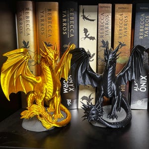 May include: Three dragon figurines standing in a row. The left dragon is metallic gold, the center dragon is matte black with a spiked tail, and the right dragon is metallic bronze. They are positioned in front of several books by Rebecca Yarros.