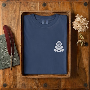 Puede incluir: Una camiseta azul marino con un gráfico blanco de una piña estilizada. La camiseta está doblada dentro de una bandeja de madera. Una pluma roja y un libro con un diseño de estrella están a la izquierda. Una flor seca está a la derecha.