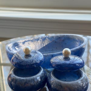May include: A set of blue decorative containers with lids, each topped with a pearl-like knob. The set includes two small round containers and a larger oval bowl, all with a marbled effect.
