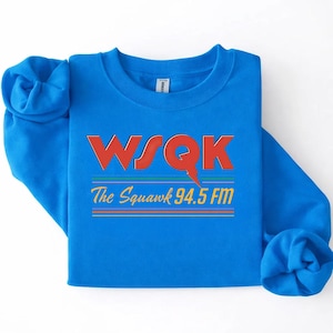 May include: A bright blue sweatshirt with the text "WSQK The Squawk 94.5 FM" in red, orange, and green. The design includes a lightning bolt graphic. The sweatshirt is folded.