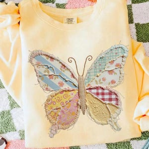 Puede incluir: Una sudadera amarilla clara con un gran aplique de mariposa. Las alas de la mariposa están hechas de varias telas estampadas, incluyendo rayas, estampados florales y cuadros. La mariposa tiene un cuerpo y antenas de color beige.