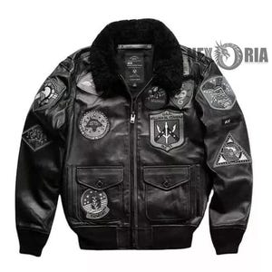May include: Black leather bomber jacket with a faux fur collar and various embroidered patches. The jacket has a zip-up front, two front pockets, and ribbed cuffs and hem. Patches include military-themed designs and text.