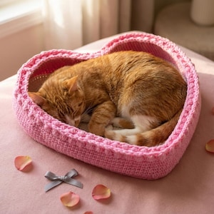 May include: A sleeping orange tabby cat curled up in a pink, heart-shaped crochet pet bed. The bed has a textured border and is surrounded by rose petals and a gray bow on a pink surface.