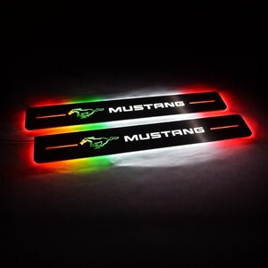 Puede incluir: Dos placas de umbral de puerta Mustang negras iluminadas con un logotipo de caballo corriendo y la palabra "MUSTANG" en blanco. Las placas tienen un degradado de iluminación LED roja, verde y blanca.