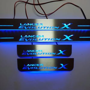 Könnte beinhalten: Satz von vier Edelstahl-Autotürschwellerschutzleisten mit blauer LED-Beleuchtung. Der Text "Lancer Evolution X" leuchtet blau. Drähte ragen von der Oberseite der Schutzleisten heraus.