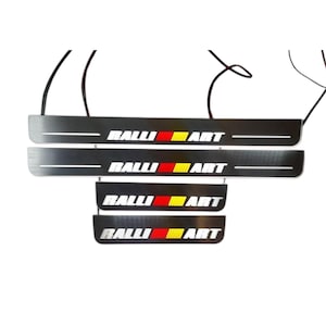 Puede incluir: Protectores de umbral de puerta de coche de metal negro con el texto "RALLI ART" en blanco, rojo y amarillo. Los protectores tienen un acabado de metal cepillado y están diseñados para proteger los umbrales de las puertas de arañazos y daños.