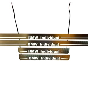 May include: Brushed metal car door sill plates with the text "BMW Individual" in a bold, sans-serif font. The plates are rectangular and have a metallic finish. Black wires are visible at the top.