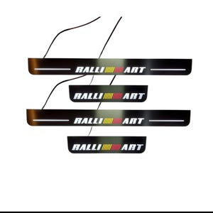 Puede incluir: Cuatro protectores de umbral de puerta de coche negros con el texto "RALLI ART" en blanco, amarillo y rojo. Cada protector tiene una barra de luz horizontal blanca. Los protectores están conectados por cables negros.