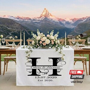 May include: A white table runner with a black monogram, the names "Olivia & Harries", and the date "13.07.2029" is set for a wedding. The table is set with flowers, candles, and glassware, with a mountain backdrop.
