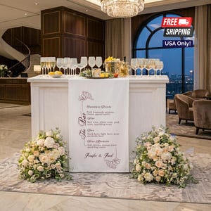 May include: A white wooden bar counter draped with a long runner featuring the text The Bar, Signature Drinks, and the names Jenifer & Jack. The counter is topped with numerous clear glass stems and two large floral arrangements sit at the base.
