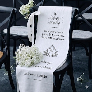 May include: A white fabric memorial sign draped over a black chair, featuring the text "in loving memory," "Your presence is gone, but your love stays with us always," "Always in our Hearts," and "Jackson Franklin" with a candle illustration.