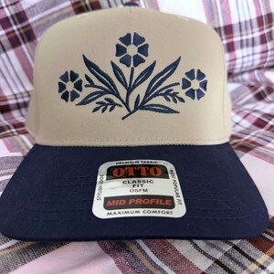 May include: A beige and navy blue baseball cap with a floral design embroidered on the front. The cap has a flat brim and a mid-profile fit. The cap has a tag that says "OTTO" and "Classic Fit".