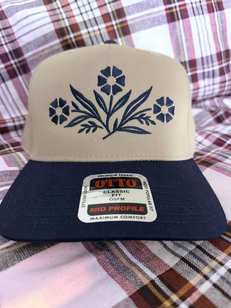 May include: A beige and navy blue baseball cap with a floral design embroidered on the front. The cap has a flat brim and a mid-profile fit. The cap has a tag that says "OTTO" and "Classic Fit".