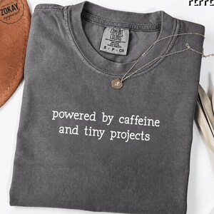 May include: A charcoal gray short-sleeve t-shirt with a crew neckline. The shirt features the white printed text "powered by caffeine and tiny projects" across the chest. A thin silver chain necklace with a small circular pendant rests on the collar.