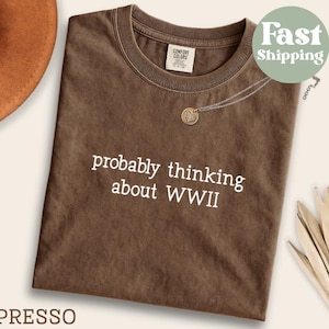 May include: Espresso-colored t-shirt with the phrase "probably thinking about WWII" in white. The shirt has a crew neck and a small tag on the upper back. A gold pendant necklace is draped on the shirt. A brown hat is visible in the upper left corner.