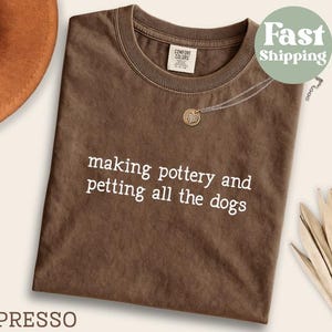 May include: Espresso-colored t-shirt with the text "making pottery and petting all the dogs" in white. The shirt has a crew neck and a small tag on the upper left. A gold pendant necklace and a brown hat are also visible.