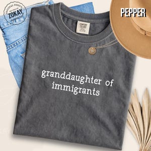 May include: A charcoal grey short-sleeve t-shirt with the white text "granddaughter of immigrants" printed on the front. The shirt features a crew neckline and a label inside the collar. The garment is displayed alongside blue denim fabric.