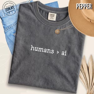 May include: A charcoal gray crew neck t-shirt featuring the white text humans > ai printed on the front. The shirt has a Comfort Colors label at the inner neckline and a circular Zokay Apparel logo stamp in the upper left corner of the image.