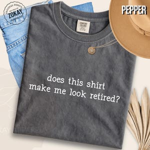 May include: A charcoal gray short-sleeve t-shirt with the white text "does this shirt make me look retired?" printed on the front. The shirt features a crew neckline and a Comfort Colors brand label on the inside collar area.